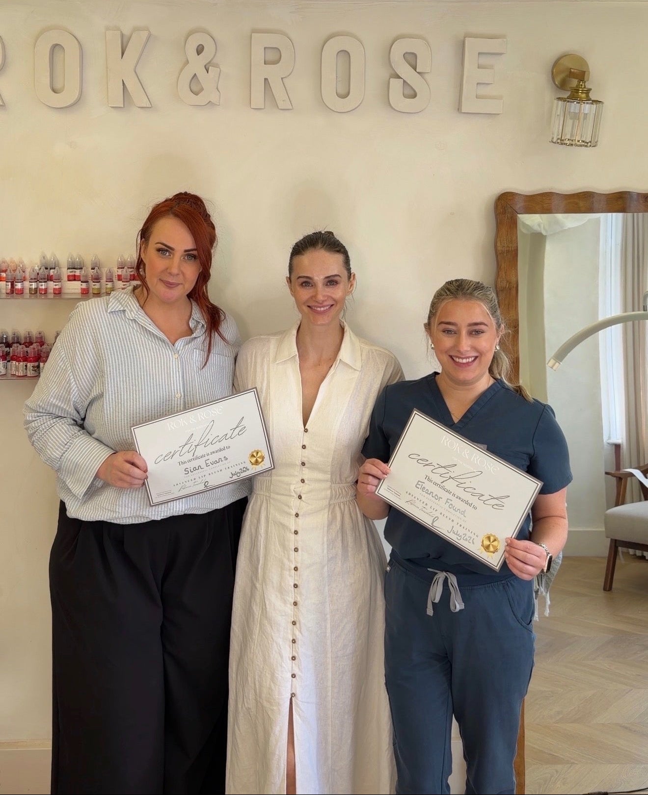 Three women standing together in a spa or treatment center, each holding a certificate. The background features a cream-colored wall with 'ROK & ROSE' in large letters, draped white fabric overhead, and a mirror on the right. Lip blush training course. Lip blush training centre Manchester Cheshire. 